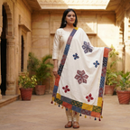 Woman holding a traditional embroidered applique dupatta in an outdoor setting with architectural elements and plants.