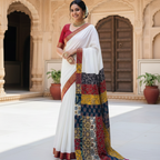 Woman in a traditional saree with a colorful pattern draped over her shoulder, standing in front of an architectural background.