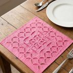 Pink placemat with cut-out pattern on a wooden table with silverware and a plate.